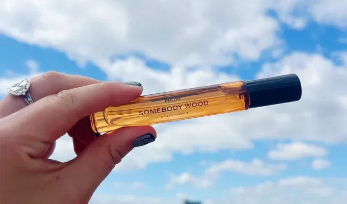 phlur-fragrance-review: A white hand adorned with rings and painted nails holds up a light orange travel-sized bottle of perfume. It’s label says Somebody Wood. Original photo.' title='I Tried All 22 Phlur Fragrances and Here’s How I’m Ranking Them (with Original Photos)