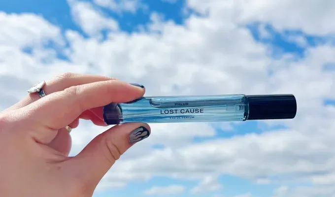phlur-fragrance-review: A white hand adorned with rings and painted nails holds up a blue travel-sized bottle of perfume. It’s label says Lost Cause. Original photo.' title='I Tried All 22 Phlur Fragrances and Here’s How I’m Ranking Them (with Original Photos)