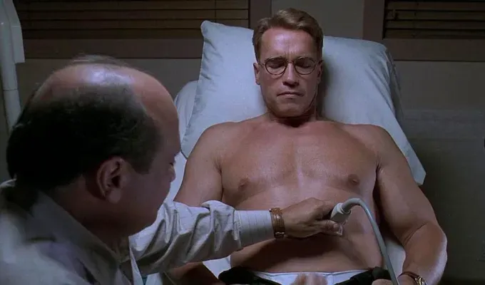 Movies About Pregnancy - A shirtless Arnold Schwarzenegger sits on a hospital bed while a male doctor does an ultrasound on his belly.' fetchpriority='auto' title='25 Movies About Pregnancy That Will Make You Laugh Cry and Give You All the Feels (And Not Because of Hormones)
