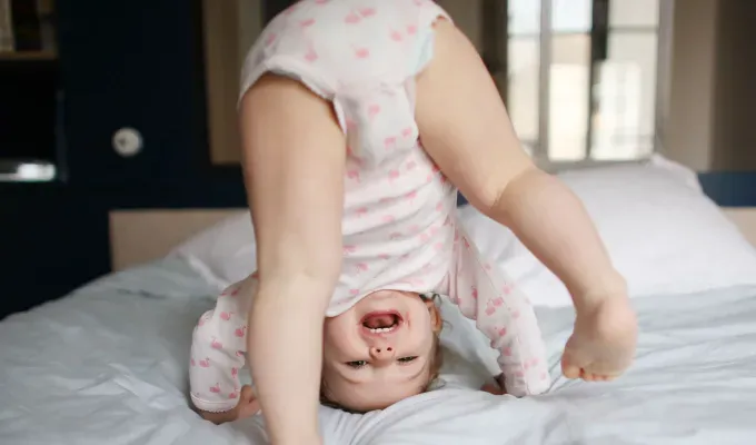 baby-names-that-mean-miracle: A young baby girl attempts a cartwheel on a bed. She peeks at the camera through her legs. She wears a onesie.' fetchpriority='auto' title='50 Baby Names That Mean Miracle