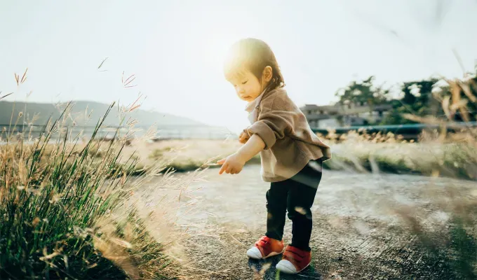 names-that-mean-summer: Curious toddler girl picking and peering at grass in the park. She wears a brown shirt dark pants and red shoes.' fetchpriority='auto' title='60 Names That Mean Summer to Celebrate Your Baby’s Sunny Spirit