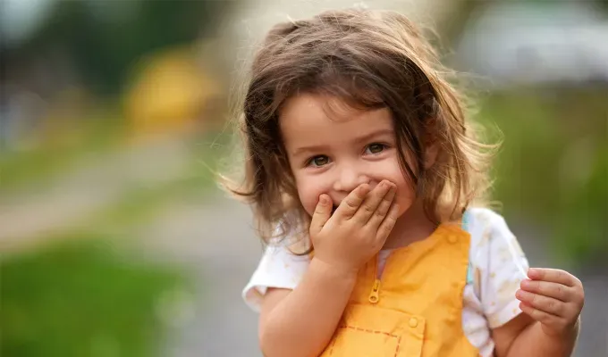 names-that-mean-summer: Little girl with brown hair outdoors covering her mouth laughing. She wears a yellow and white top.' fetchpriority='auto' title='60 Names That Mean Summer to Celebrate Your Baby’s Sunny Spirit