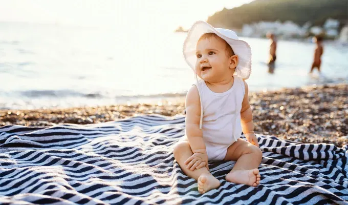 names-that-mean-summer: Baby girl sitting on towel at the beach in summer. She wears a white t-shirt diaper and sun hat.' fetchpriority='auto' title='60 Names That Mean Summer to Celebrate Your Baby’s Sunny Spirit