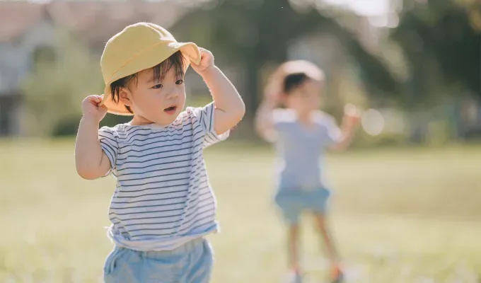 names-that-mean-summer: A young Asian boy wearing yellow hat and black and white striped top at public park in the morning. Behind him another child is blurred in the background.' fetchpriority='auto' title='60 Names That Mean Summer to Celebrate Your Baby’s Sunny Spirit