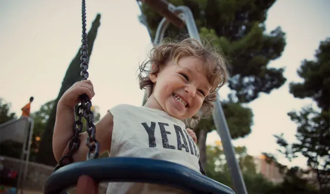 names-that-mean-summer: Toddler at the swing laughing. The angle of the shot is from below and the child wears a white t-shirt that says Yeah! on it. He has brown hair and swings in front of some trees.' fetchpriority='auto' title='60 Names That Mean Summer to Celebrate Your Baby’s Sunny Spirit