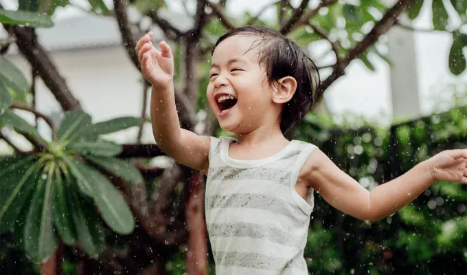 names-that-mean-summer: A young Asian boy with a green and white striped shirt on plays in a sprinkler in front of some bushes. He is laughing.' fetchpriority='auto' title='60 Names That Mean Summer to Celebrate Your Baby’s Sunny Spirit