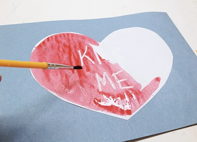 52 Fun and Easy Valentine’s Day Crafts for Kids - A paper craft that features a blue construction paper sheet a smaller heart-shaped white sheet of paper with the words ’Kiss Me’ written in white crayon. Someone is painting over the white heart with red watercolor to reveal the ’Kiss Me’ text.' title='70 Fun and Easy Valentine’s Day Crafts for Kids
