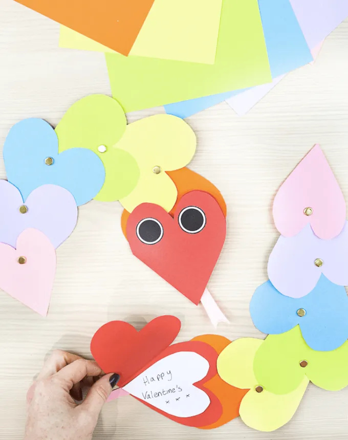52 Fun and Easy Valentine’s Day Crafts for Kids - A rainbow snake Valentine’s craft made with various heart shaped pieces of paper affixed by pins. The first heart has eyes on it as well as a tongue sticking out from it and a Valentine note written inside the head.' title='70 Fun and Easy Valentine’s Day Crafts for Kids