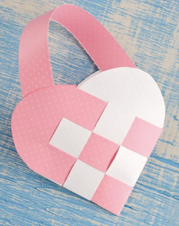 52 Fun and Easy Valentine’s Day Crafts for Kids - A pink and white woven paper Valentine’s basket. It’s featured on a blue and white background.' title='70 Fun and Easy Valentine’s Day Crafts for Kids