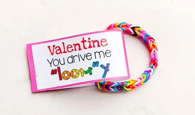 52 Fun and Easy Valentine’s Day Crafts for Kids - A rainbow colored Loom bracelet made of rubber bands rests on a white surface. Attached to the bracelet is a tag that reads ’Valentine You drive me loomy.’' fetchpriority='auto' title='70 Fun and Easy Valentine’s Day Crafts for Kids