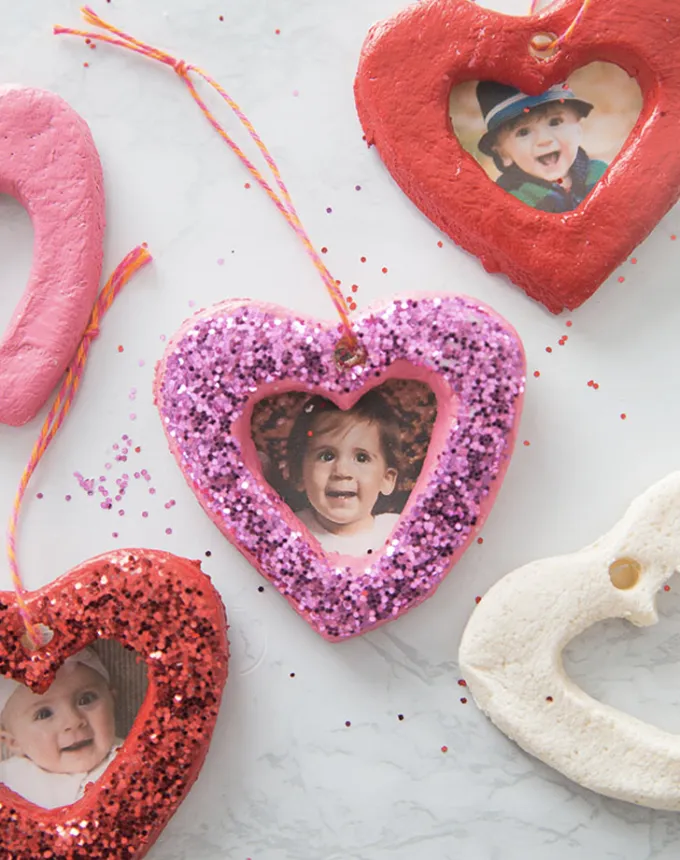 52 Fun and Easy Valentine’s Day Crafts for Kids - Salt dough ornaments covered in pink and red paint and glitter are affixed with photos of children and hung on strings for decorative use.' title='70 Fun and Easy Valentine’s Day Crafts for Kids