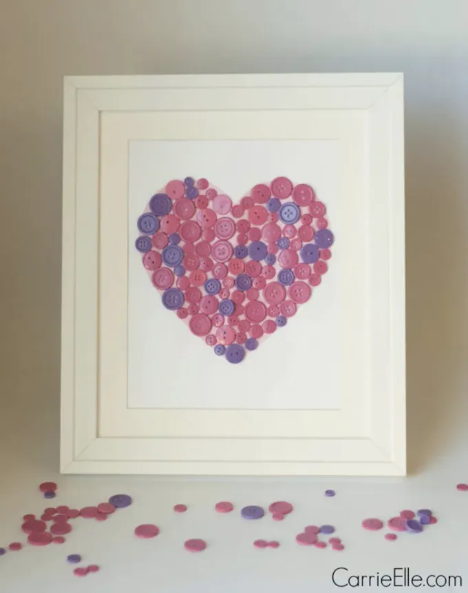 52 Fun and Easy Valentine’s Day Crafts for Kids - A decorative piece of art that features purple and pink buttons arranged in the shape of a heart. It’s framed in a white frame with a white mounting. There are buttons scattered on the white surface that the photo sits on.' title='70 Fun and Easy Valentine’s Day Crafts for Kids