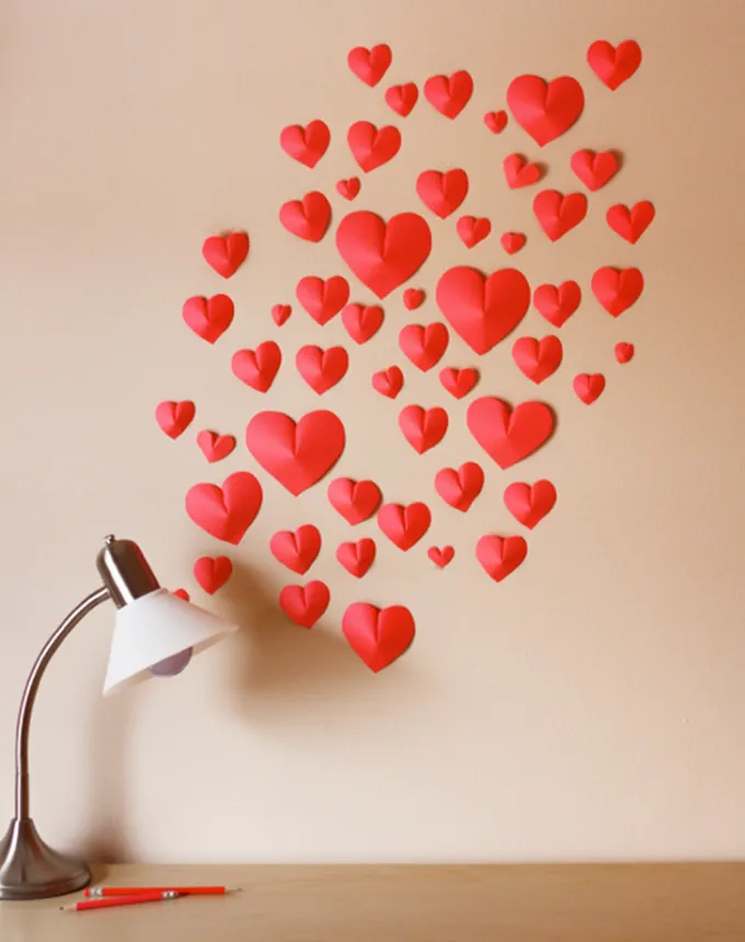 52 Fun and Easy Valentine’s Day Crafts for Kids - An array of different sized red origami hearts that are adhered to a wall above a desk with a table lamp.' title='70 Fun and Easy Valentine’s Day Crafts for Kids