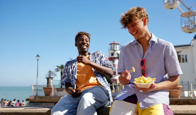 gen-z-slang-terms: Two teens sit on a boardwalk at the beach laughing and eating food together.' fetchpriority='auto' title='From ‘Basic’ to ‘Boujee’ Here Are 29 Gen Z Slang Terms To Keep in Your Back Pocket