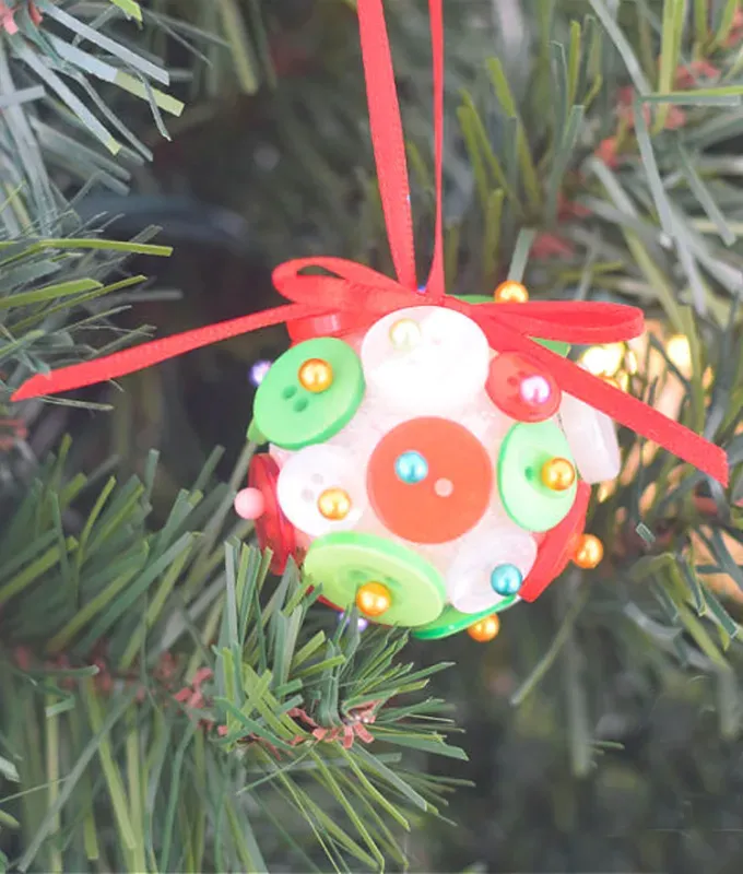 Christmas Crafts for Toddlers - A white ornament covered in red and green buttons hangs from a red ribbon on a pine tree.' title='35 Christmas Crafts for Toddlers That They Can Actually Do