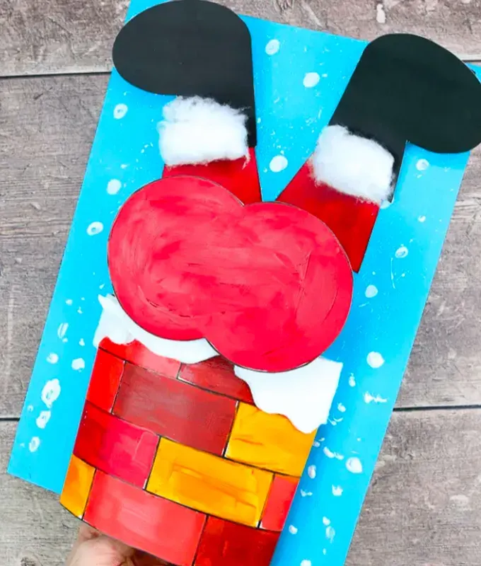 Christmas Crafts for Toddlers - A paper craft made to look like Santa’s stuck inside of a chimney. The only visible parts of Santa are his butt and legs. Santa’s wearing red pants cotton ball fluff for socks and black shoes. He is stuck in a red and yellow brick chimney. The background of the craft shows a bright blue sky with snowflakes.' title='35 Christmas Crafts for Toddlers That They Can Actually Do