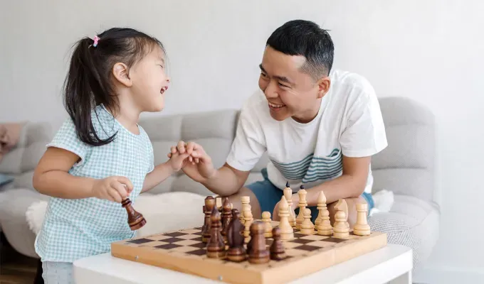 father-daughter-date-ideas: young Asian father playing chess with little daughter on sofa in living room looking at each other and laughing. The daughter wears a light blue and white striped shirt and matching pants and the dad wears a white t-shirt and jeans.' fetchpriority='auto' title='30 Father-Daughter Date Ideas for a Memorable Bonding Experience