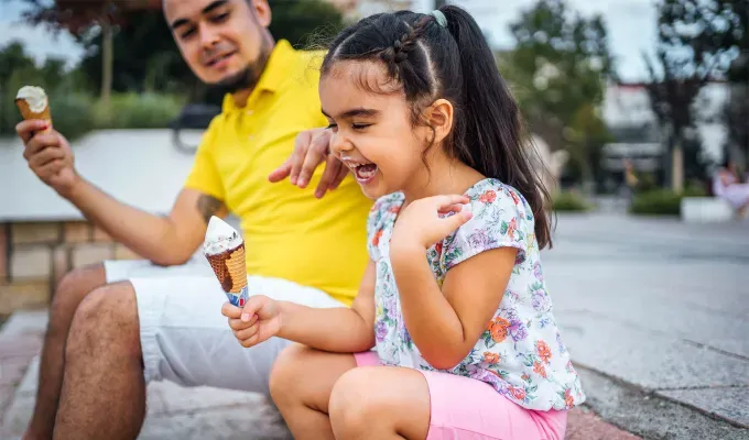 father-daughter-date-ideas: Young father with daughter eating ice cream in the city. They seem to be of South Asian descent. The father wears a yellow shirt and white shorts and the daughter wears a floral top and pink shorts. She has long dark hair in a ponytail that starts as a braid. She laughs and has ice cream on her lips.' fetchpriority='auto' title='30 Father-Daughter Date Ideas for a Memorable Bonding Experience