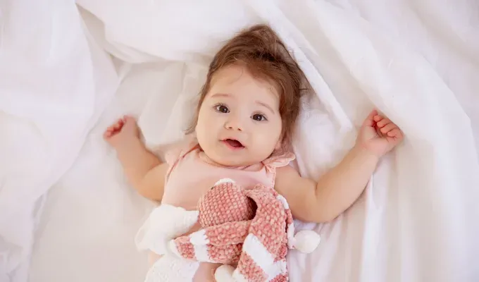 baby-names-that-mean-love: Baby girl in pink bodysuit lying on the bed at home looking at the camera top view.' fetchpriority='auto' title='75 Baby Names That Mean Love