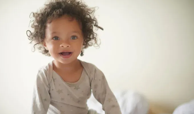 baby-names-that-mean-love: A photo of a young girl in her pajamas sitting on a bed. She seems to be mixed race and has brown curly hair and light eyes. Her pajamas are a light gray with stars.' fetchpriority='auto' title='75 Baby Names That Mean Love