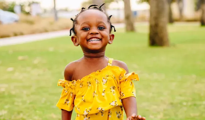 baby-names-that-mean-love: A young Black girl with hair twists wears a yellow romper while running around outside in a lush green park. She is smiling.' fetchpriority='auto' title='75 Baby Names That Mean Love