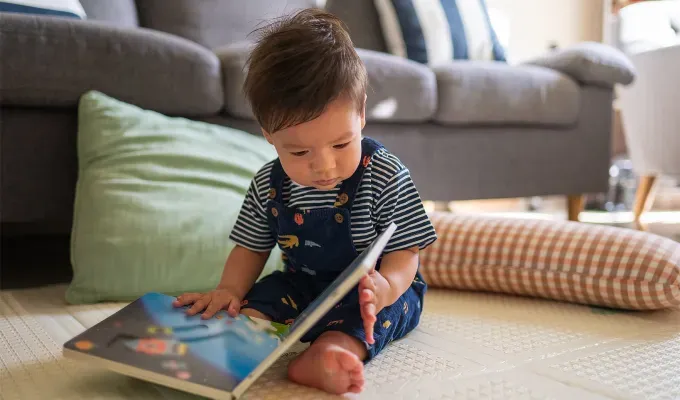 baby-names-that-mean-love: Baby boy looking at a book in the living room sitting on a baby safe soft play mat on the floor at home. He seems to be mixed race with brown hair and brown eyes. He wears a striped shirt and overalls with little designs.' fetchpriority='auto' title='75 Baby Names That Mean Love