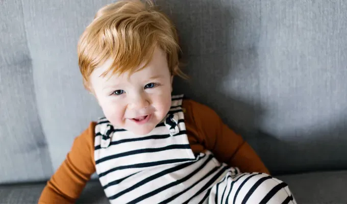 baby-names-that-mean-love: A young white boy with ginger hair sits on a couch and smiles at the camera. He wears an orange long sleeved top and black and white striped overalls. The couch is gray.' fetchpriority='auto' title='75 Baby Names That Mean Love