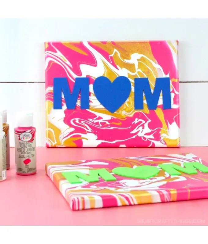 mothers-day-crafts: Two swirled paintings with pink orange and white paint and letters that spell out Mom with a heart as the O. They sit on a pink table with bottles of paint.' title='45 Mother’s Day Crafts for Kids That She’ll Actually Want to Keep