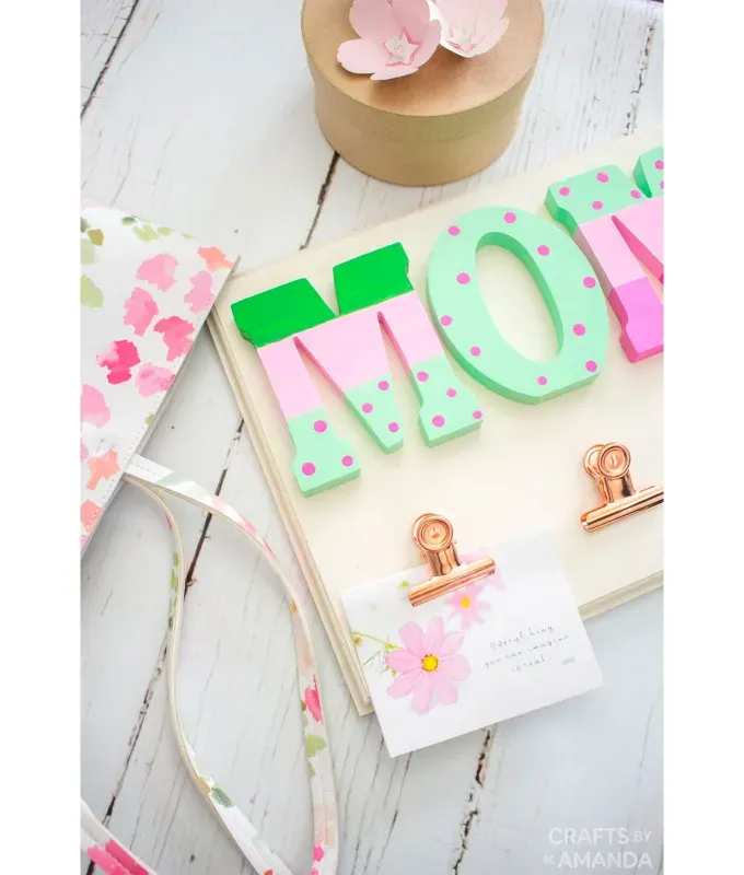 mothers-day-crafts: A wooden sign with the word Mom spelled out in green and pink letters. There are metal clips to hold photos and some extra decoration surrounding it. It sits on a white wooden surface.' title='45 Mother’s Day Crafts for Kids That She’ll Actually Want to Keep