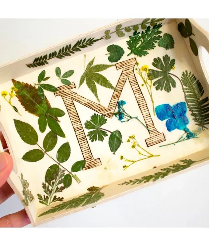 mothers-day-crafts: A light wood tray with modge podged leaves and flowers stuck to it. It has an M in the center.' title='45 Mother’s Day Crafts for Kids That She’ll Actually Want to Keep