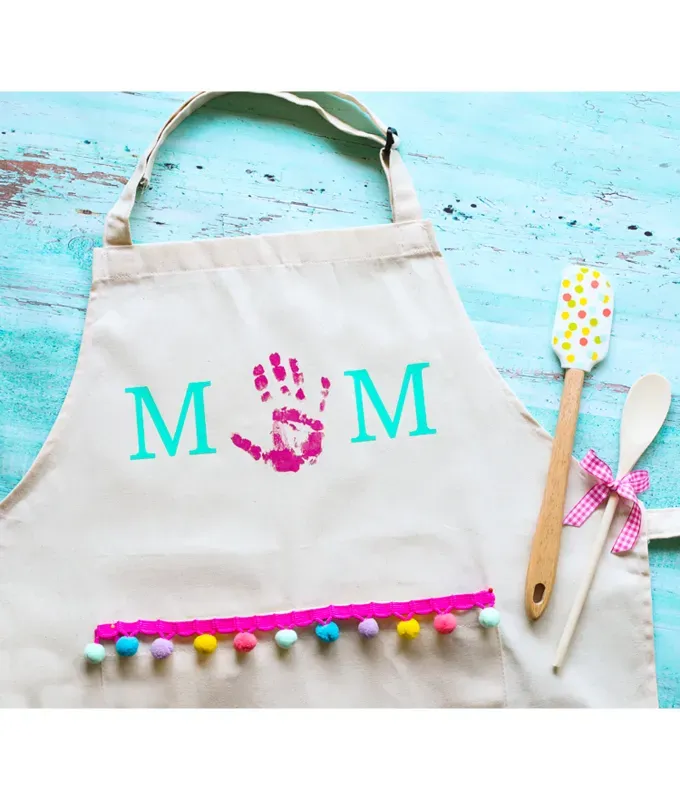 mothers-day-crafts: A white apron with the word Mom spelled out on it. The M’s are blue and the O is a blue handprint. It has a colorful pop pom trim and lays on a blue wood surface with spatulas next to it.' title='45 Mother’s Day Crafts for Kids That She’ll Actually Want to Keep