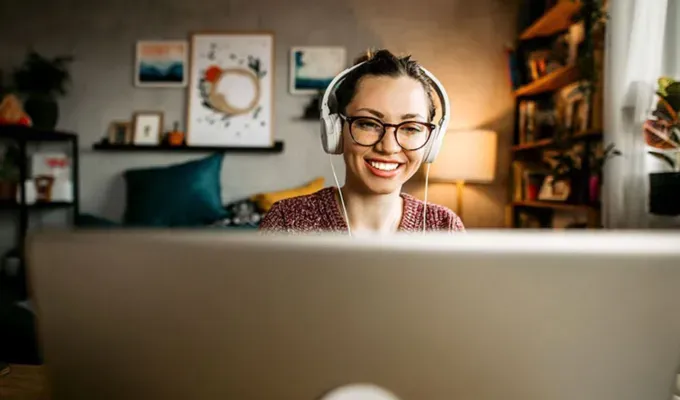 things-to-do-when-you’re-bored: A young white woman with brown hair wears glasses and a headset while smiling at her computer screen.' fetchpriority='auto' title='Wondering What to Do When You’re Bored? Here Are 105 Fun Things to Do for Any Mood