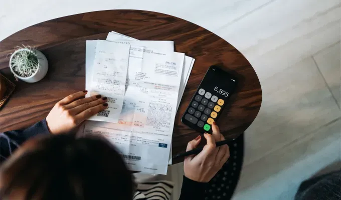 things-to-do-when-you’re-bored: A person looks over a number of papers and bills on a wood table. They are using a calculator to do math.' fetchpriority='auto' title='Wondering What to Do When You’re Bored? Here Are 105 Fun Things to Do for Any Mood