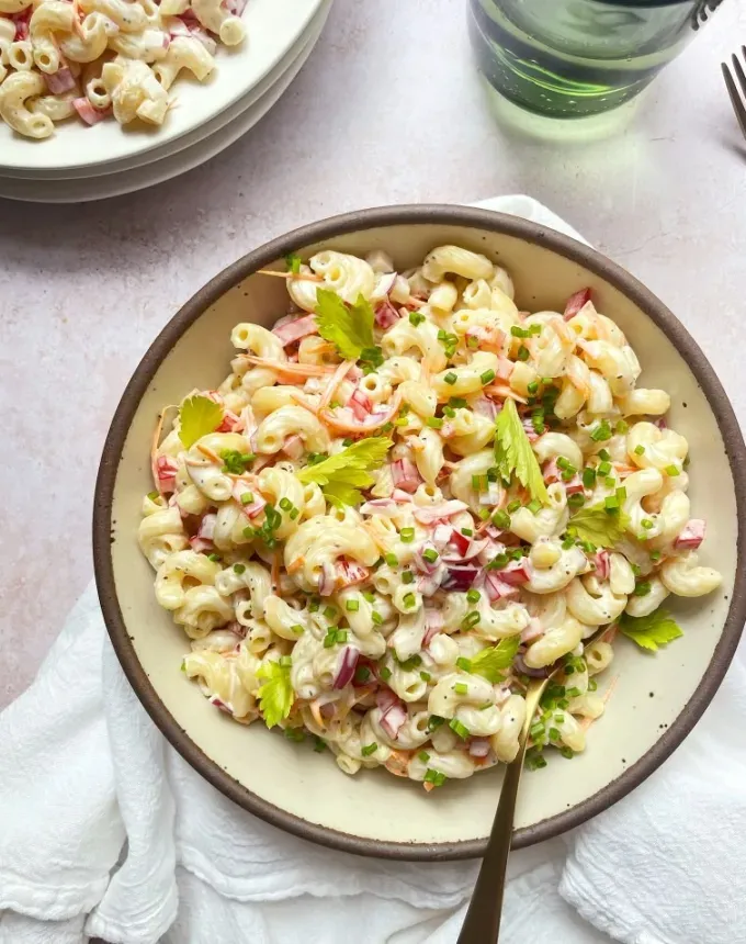big batch recipes: bowl of classic macaroni salad with a spoon it it on a table next to a glass and stack of plates' title='76 Big-Batch Dishes & Recipes to Feed a Crowd