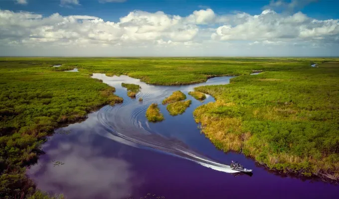 Fall Getaways in Florida - An aerial shot of a person driving a boat through the Everglades in Florida. It’s a body of water with lots of green marshes and land masses.' title='The 12 Best Fall Getaways in Florida (Because There’s Way More to See Than Just Beaches)