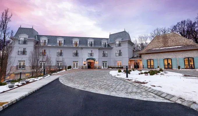 Cozy Winter Weekend Getaways from New York - The driveway leading up to the Mirbeau Inn and Spa. The building itself is ornate and gray with a smaller teal building next to it.' title='The 34 Coziest Winter Weekend Getaways from NYC in 2025