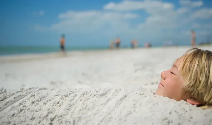 father-son-trip-ideas: A young white boy with blond hair is burried in sand up to his neck on a beach. He smiles with his eyes closed and looks up at the sky. There are clouds in the sky and people walking in the distance.' title='10 Father-Son Trip Ideas for Some Serious Bonding
