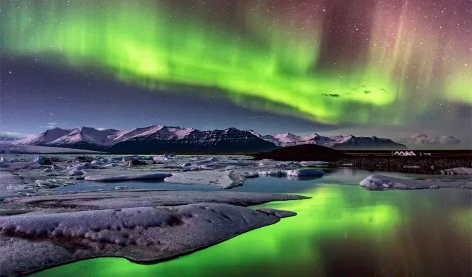 father-son-trip-ideas: A photo of the Northern Lights taken in Reykjavic Iceland. The lights are a bright green with some purplish haze. There is a reflection of the lights in a body of water surrounding the area of icy mountains.' title='10 Father-Son Trip Ideas for Some Serious Bonding
