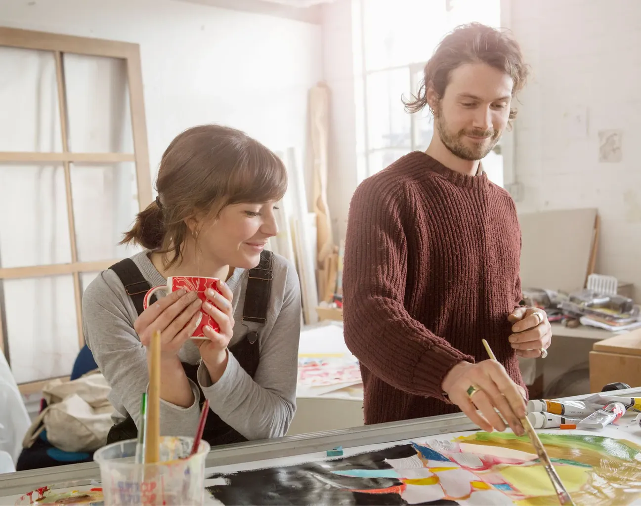 hobbies-for-couples: A man and a woman stand in an art studio at a table. The man is painting and the woman is leaning over the table holding a mug watching.' fetchpriority='high' title='47 Hobbies for Couples Looking to Connect and Have Fun