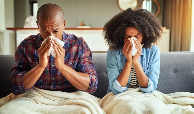 What Might Happen If You Stop Drinking Soda - A man and a woman are siting on a couch with a white blanket on their laps. They are both blowing their noses into tissues at the same time.' fetchpriority='auto' title='8 Things That Might Happen If You Stop Drinking Soda